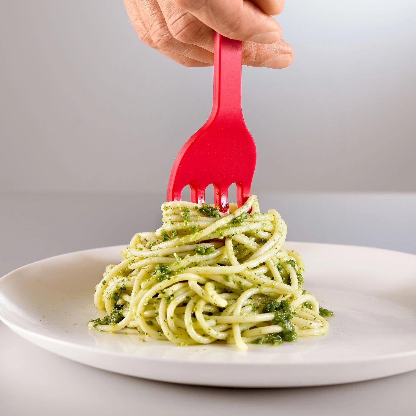 Ein roter Gabel sticht in einen Teller mit Spaghetti mit Pesto.