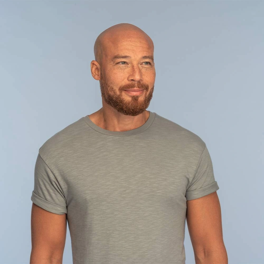 Ein Mann mit Glatze und Bart, der ein graues T-Shirt trägt, lächelt.