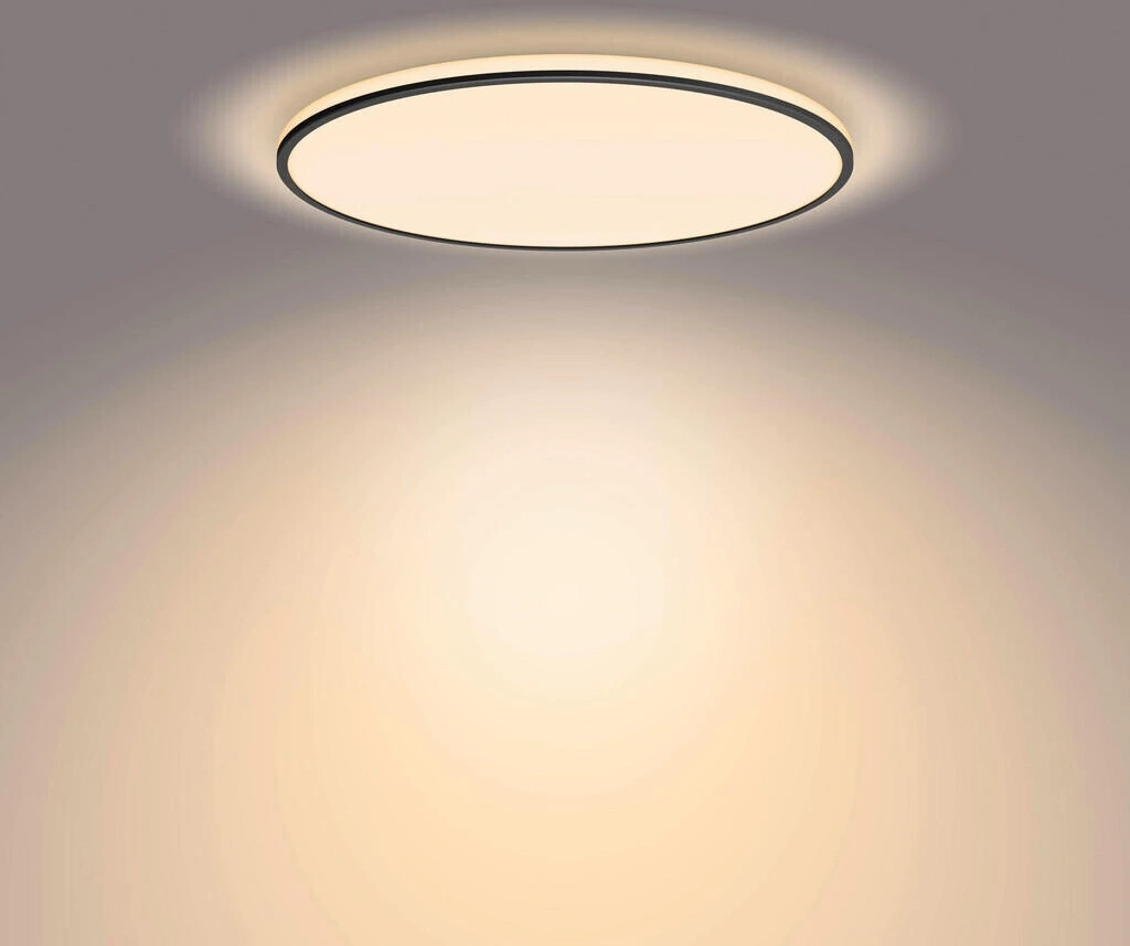 Eine moderne Deckenleuchte mit schwarzem Rand leuchtet warmes Licht auf die Wand.