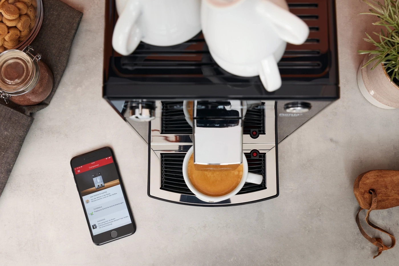 Kaffeemaschine mit Smartphone, Kaffee und Zubehör auf einer grauen Oberfläche.