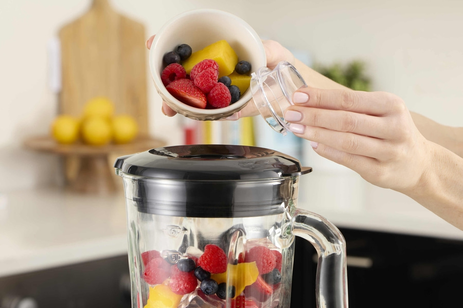 Person gießt Früchte in einen Mixer, um einen Smoothie zuzubereiten. Erdbeeren, Himbeeren, Blaubeeren und Mango.