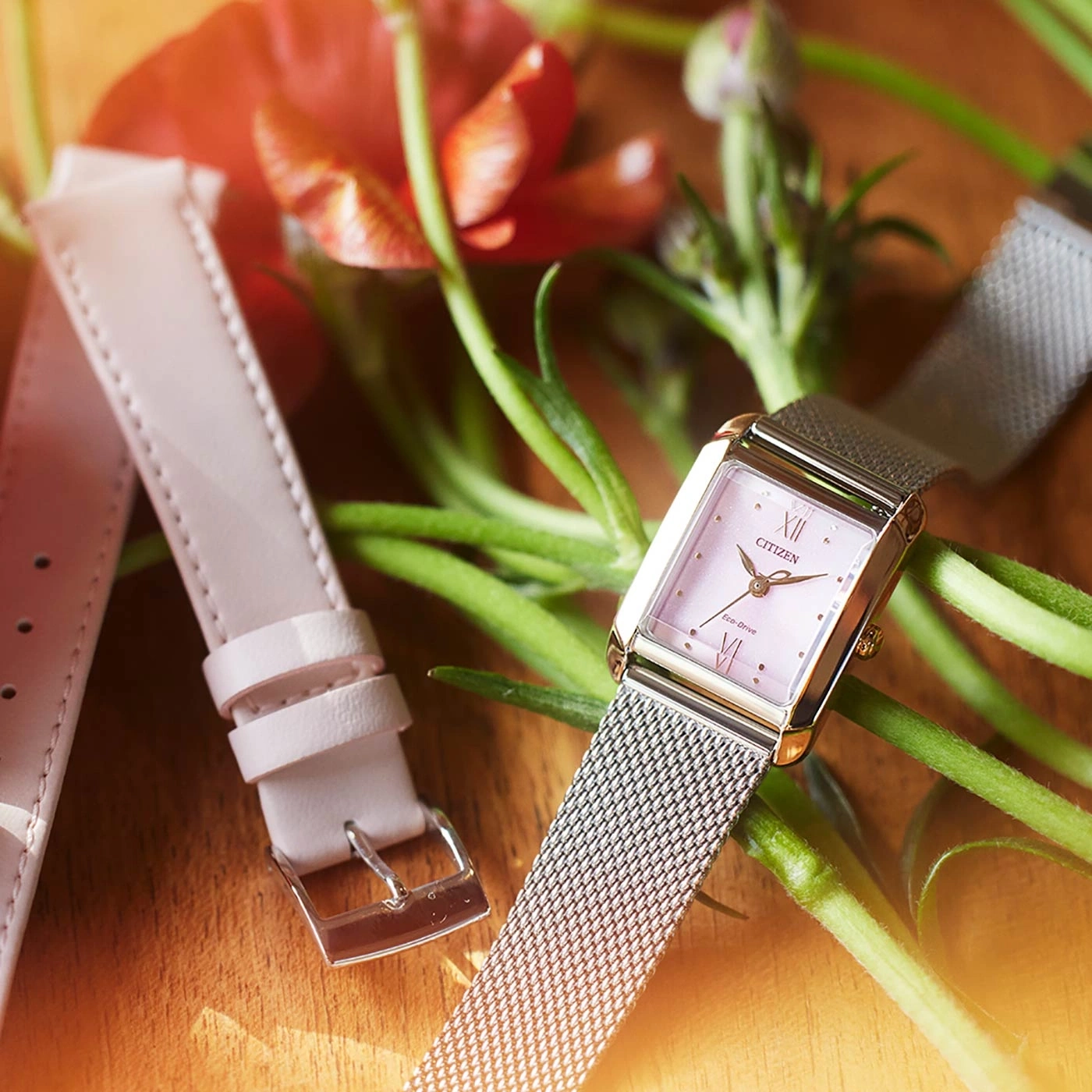 Nahaufnahme einer Citizen-Uhr mit rosa Zifferblatt und Armband, Blumen im Hintergrund