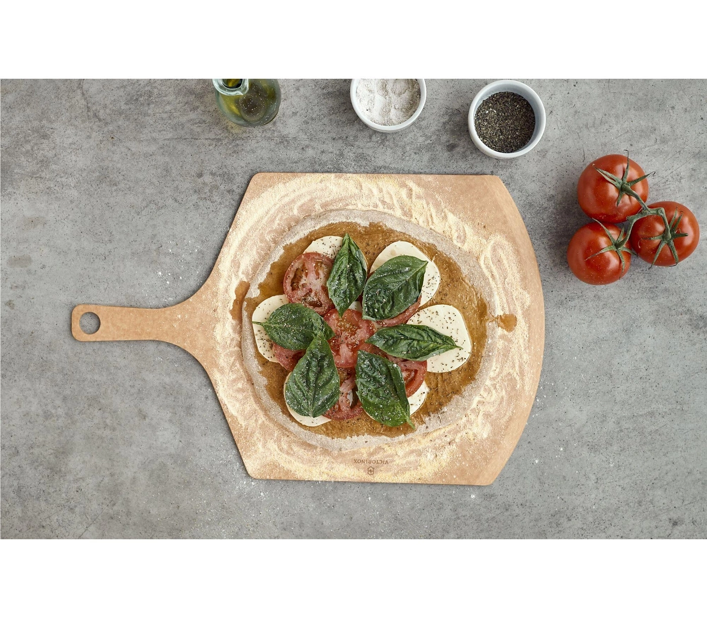 Pizza mit Tomaten, Mozzarella und Basilikum auf einem Holzbrett, daneben Zutaten