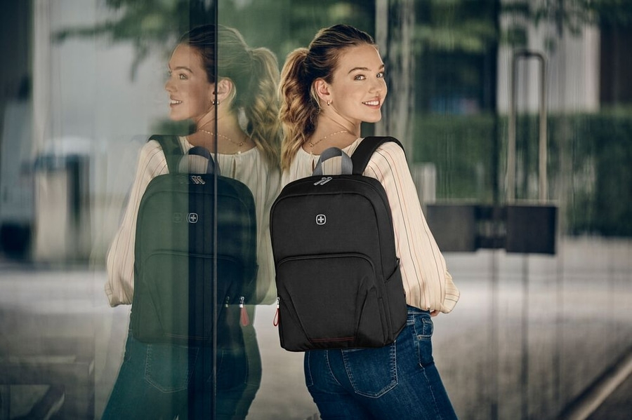 Junge Frau mit schwarzem Rucksack, die in die Kamera lächelt, vor einem Glasfenster