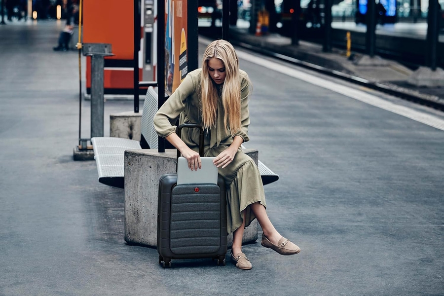Junge Frau öffnet Laptop an einem Bahnhof neben einem Koffer.