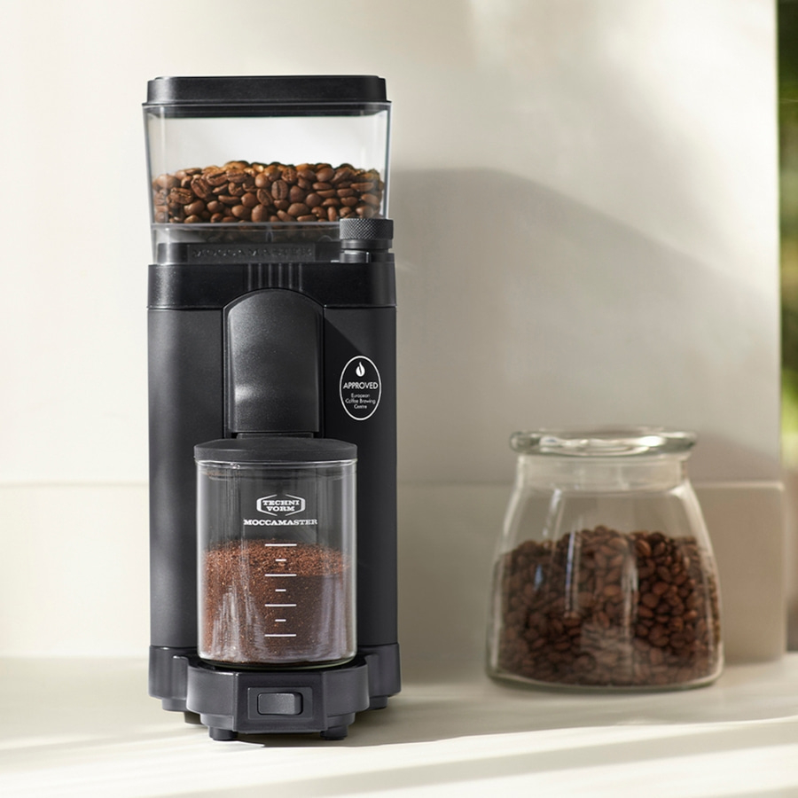 Schwarze Kaffeemühle mit Bohnen und gemahlenem Kaffee, daneben ein Glas mit Bohnen.