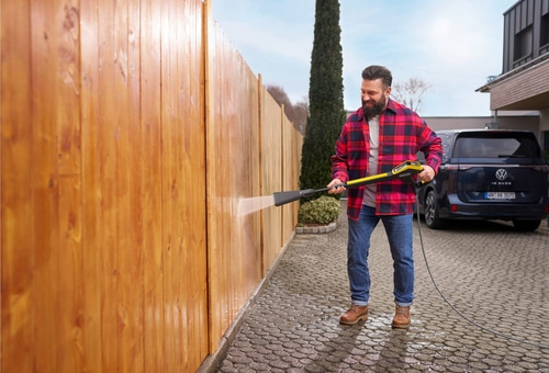 Mann reinigt Holzzaun mit Hochdruckreiniger, Auto im Hintergrund, Haus, Pflastersteine