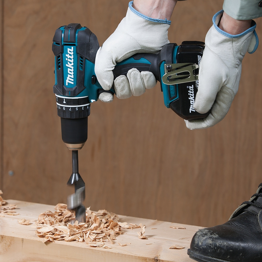 Person bohrt mit Makita-Bohrmaschine in Holz, trägt Handschuhe und Schuhe.