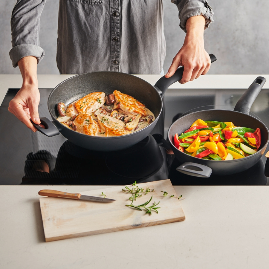 Person kocht mit zwei Pfannen, eine mit Hähnchen, die andere mit Gemüse.