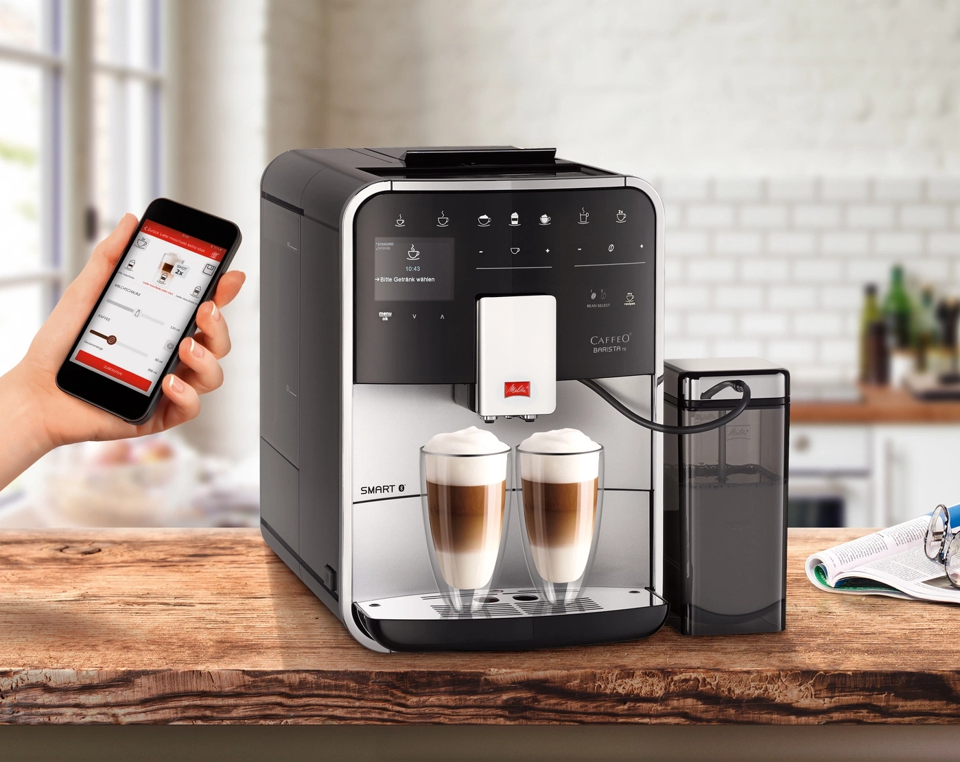 Kaffeemaschine mit Smartphone-Steuerung, zwei Gläsern Latte Macchiato und Zubehör auf einem Holztisch.