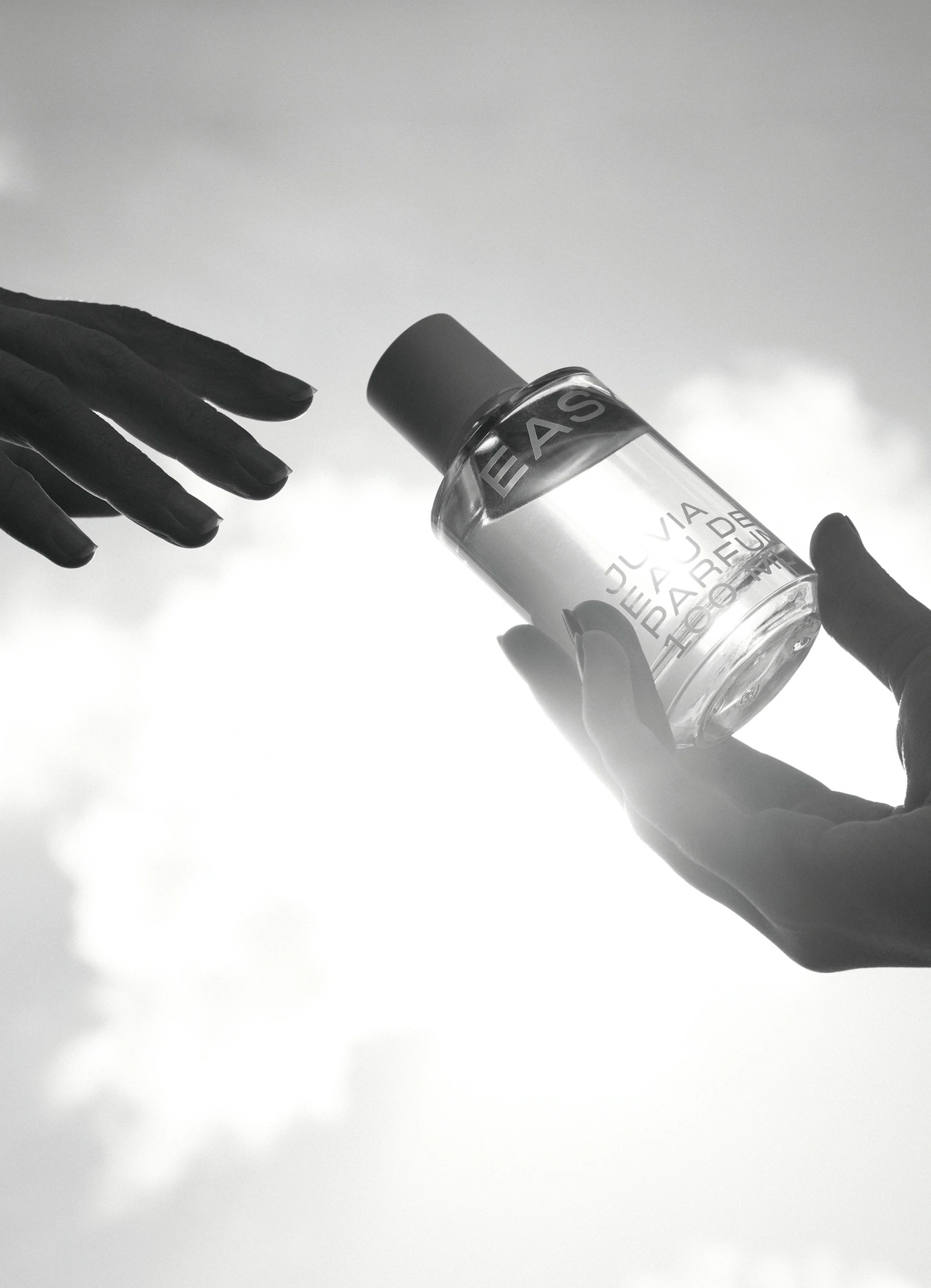 Schwarze Hände reichen sich eine Parfümflasche vor hellem Himmel.