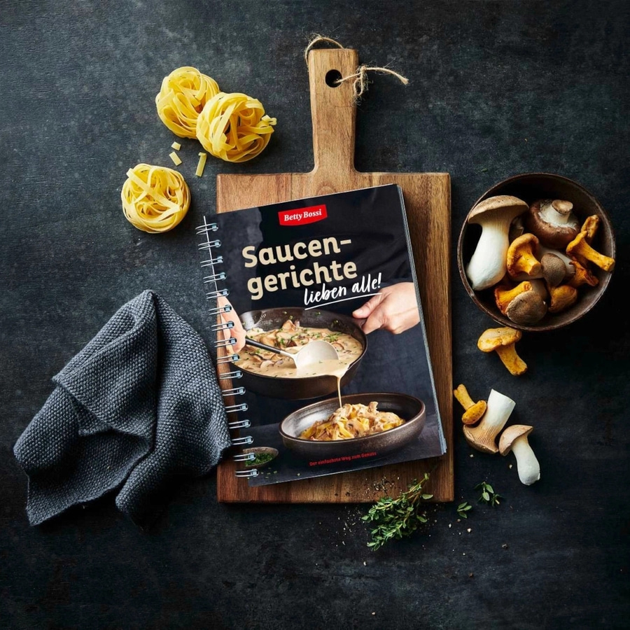 Kochbuch mit Nudeln, Pilzen und Kräutern auf einem Holzbrett