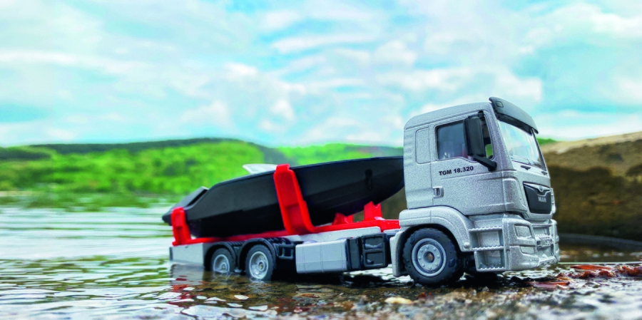 Spielzeug-LKW mit Boot im Wasser, grauer LKW, roter Halter, blauer Himmel, grüne Berge