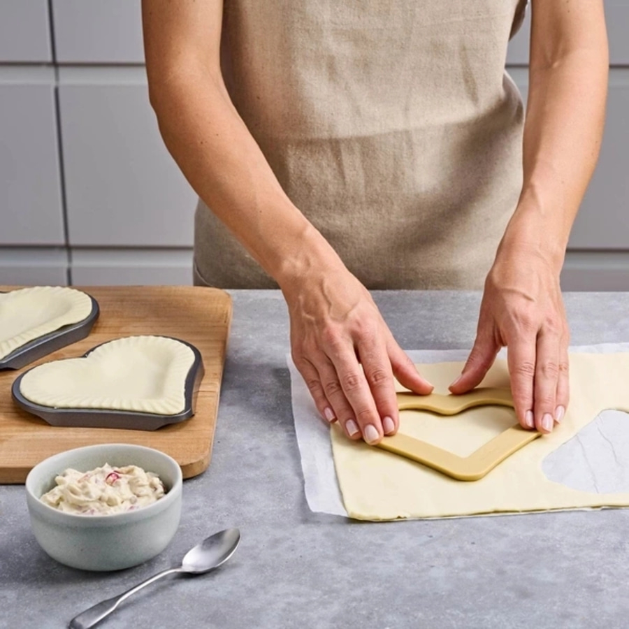 Person schneidet Teig mit Herzform aus. Herzförmige Backformen und Schüssel mit Füllung.