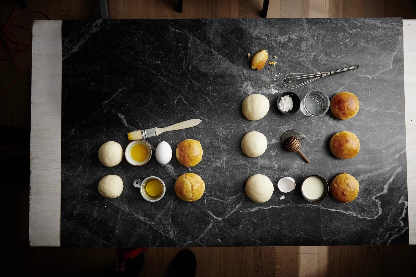 Dunkler Tisch mit Backzutaten: Teig, Eier, Öl, Honig, Wasser, Mehl, Brötchen.