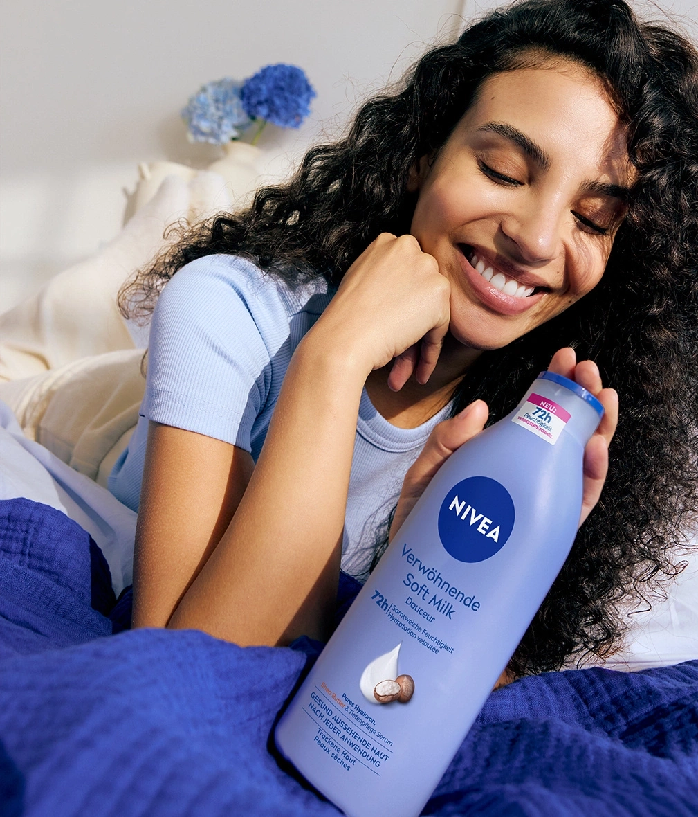Frau hält Nivea Soft Milk, lächelt, liegt im Bett, blaue Kleidung, Hautpflege