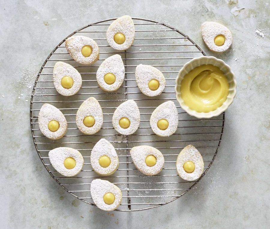 Kekse mit gelber Füllung auf einem Rost, Puderzucker, Schüssel mit Creme