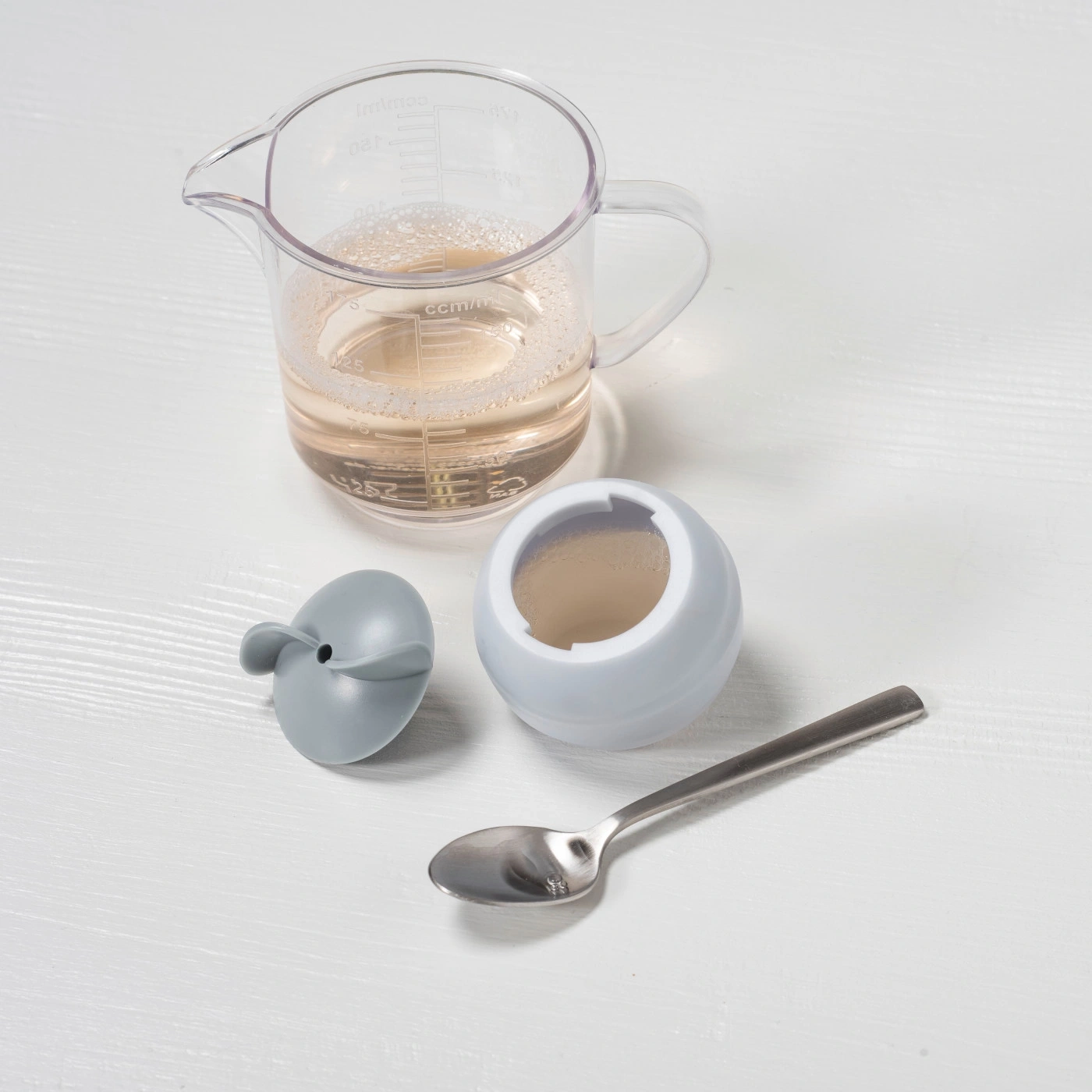 Ein Messbecher mit Flüssigkeit, ein Deckel, eine Schüssel und ein Löffel auf weißem Hintergrund.