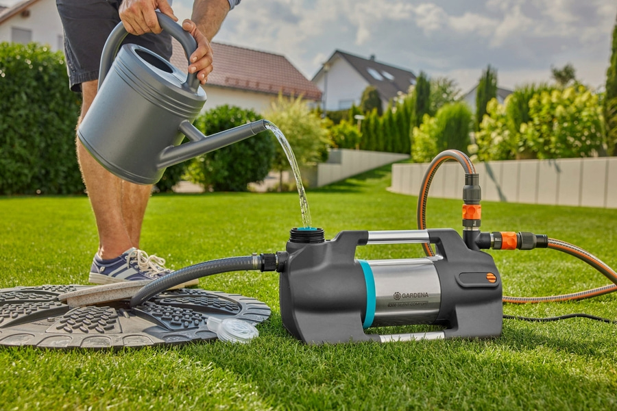 Mann gießt Wasser in eine Pumpe im Garten, Rasen, Häuser im Hintergrund