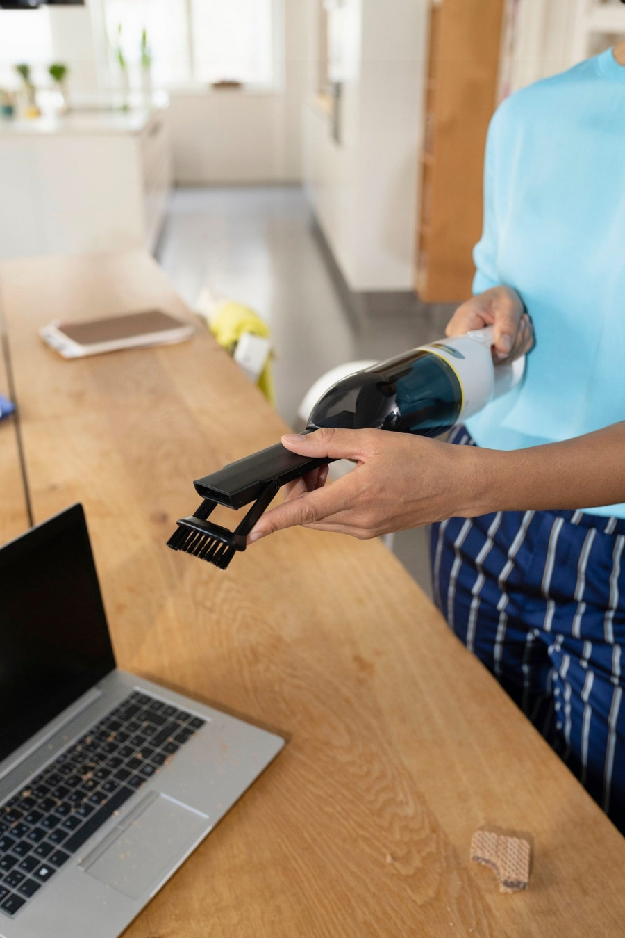 Person reinigt Laptop mit Staubsauger, auf einem Holztisch, mit Waffelresten.