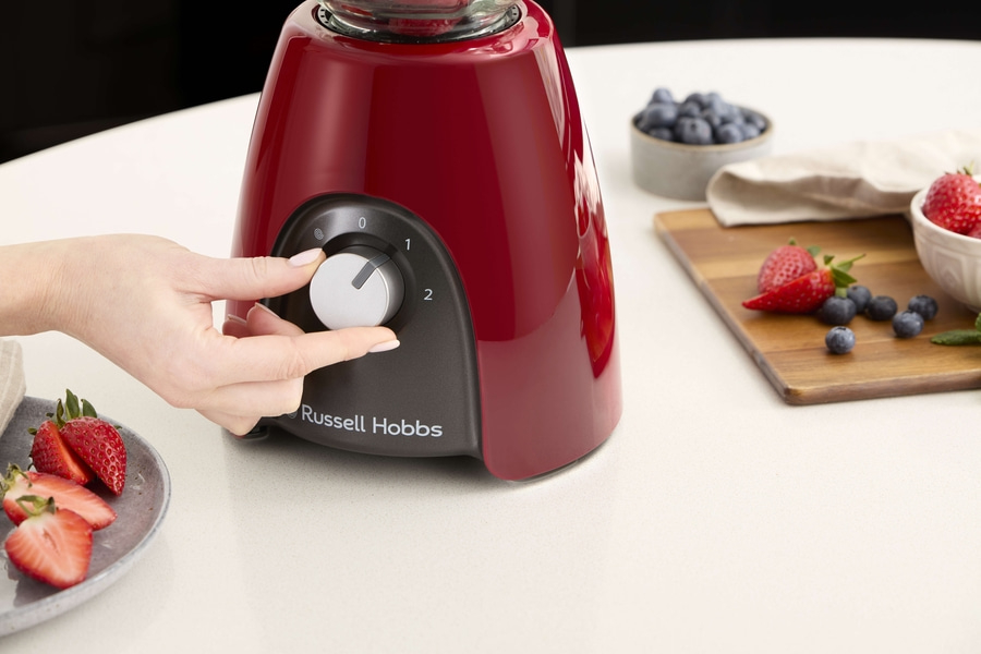 Person bedient einen roten Mixer mit Erdbeeren und Blaubeeren auf dem Tisch.