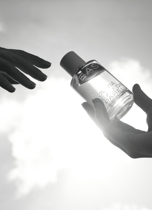 Schwarze Hände reichen sich eine Parfümflasche vor hellem Himmel.