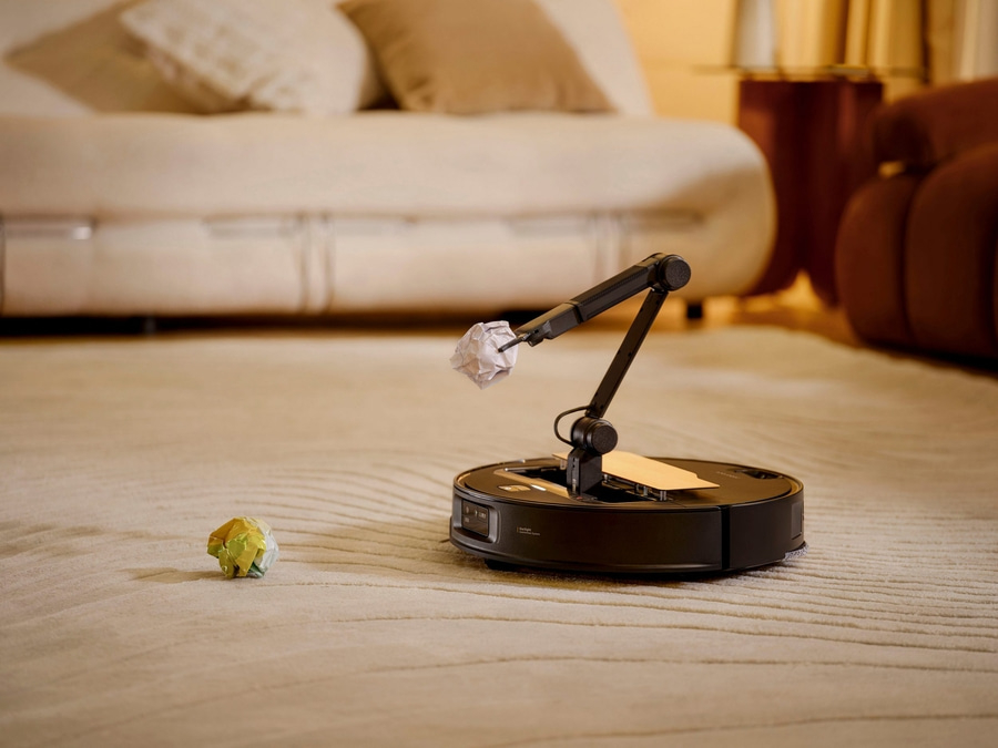 Ein Saugroboter mit Roboterarm hält ein zerknülltes Papier auf einem Teppich.