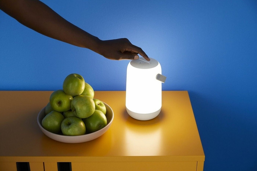 Eine Hand berührt eine Lampe neben einem Teller mit Äpfeln auf einem gelben Tisch.