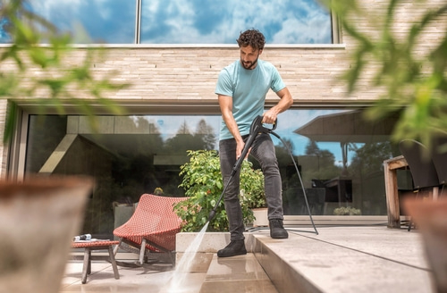Mann reinigt Terrasse mit Hochdruckreiniger. Haus im Hintergrund, sonniger Tag.