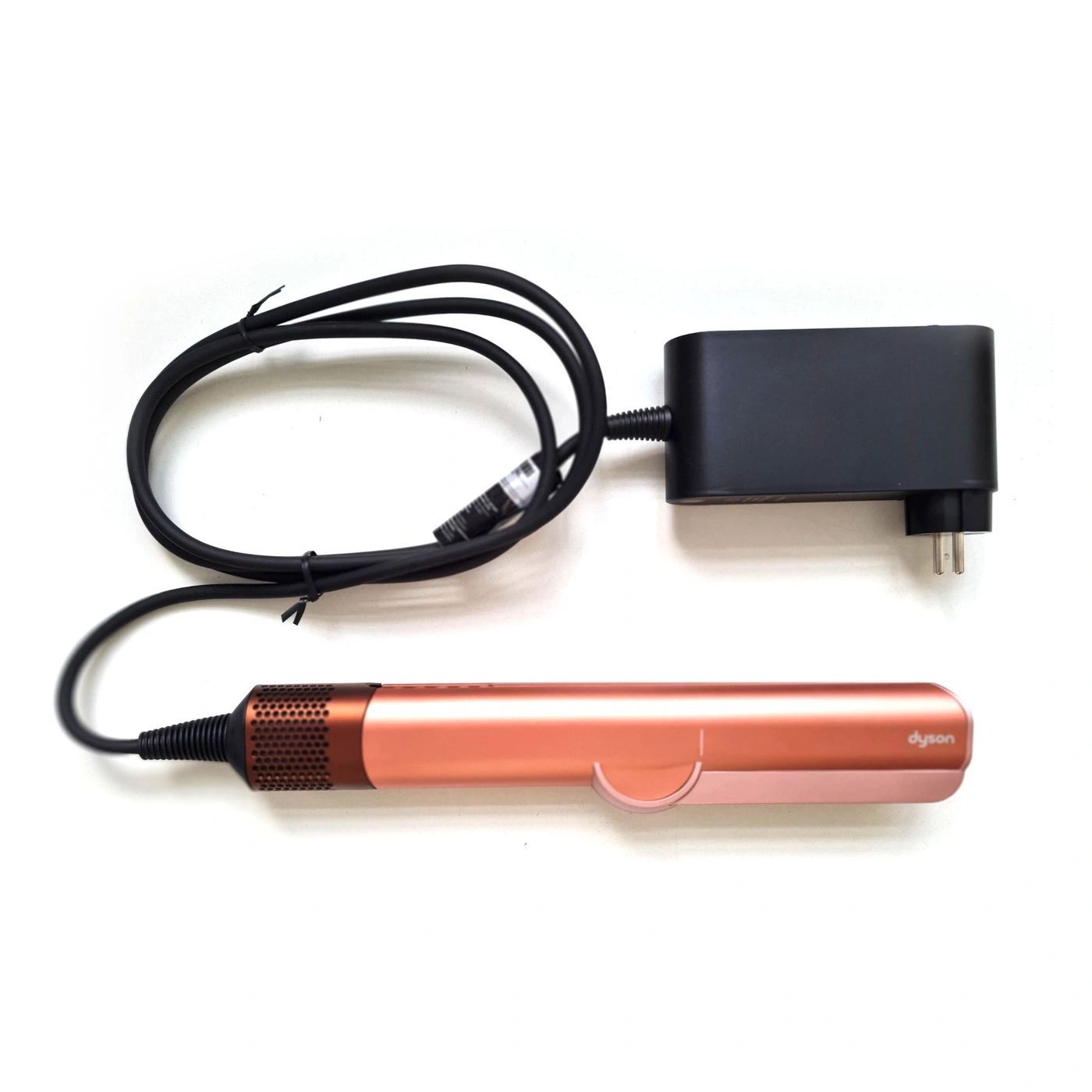 Ein roségoldener Dyson Haarglätter mit Ladekabel und Netzteil auf weißem Hintergrund.