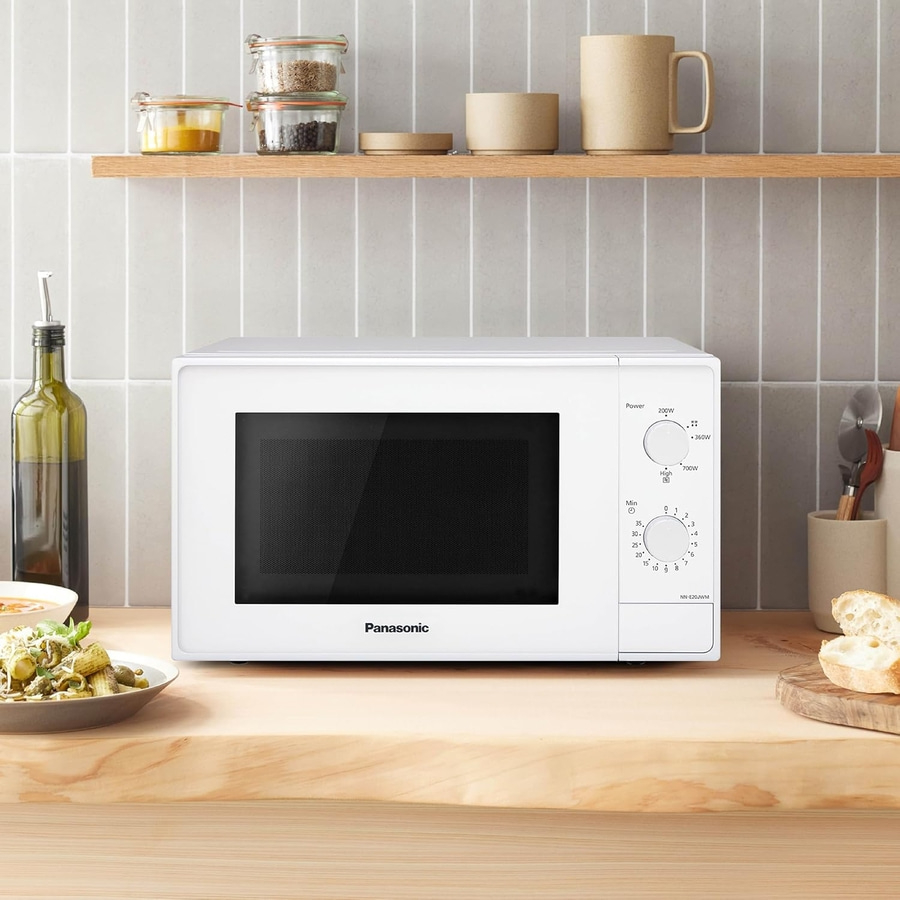 Eine weiße Panasonic Mikrowelle steht auf einer Holzoberfläche, daneben ein Teller mit Pasta und Brot.