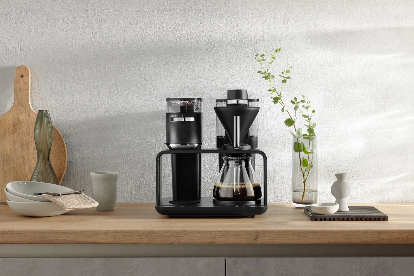 Kaffeemaschine auf einer Holzoberfläche mit Dekoration und Pflanzen vor einer weißen Wand