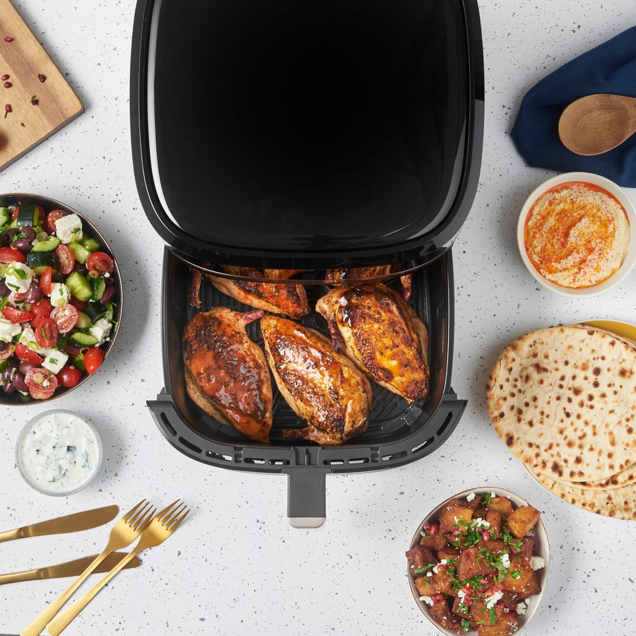 Luftfritteuse mit Hähnchen, Salat, Hummus, Brot und Beilagen auf weißem Tisch