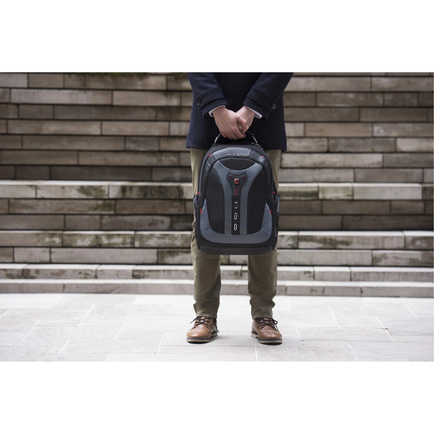 Mann hält Rucksack vor einer Steintreppe. Er trägt eine Jacke und braune Schuhe.