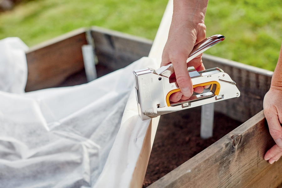 Hand benutzt Tacker, um weiße Folie an Holzrahmen zu befestigen.