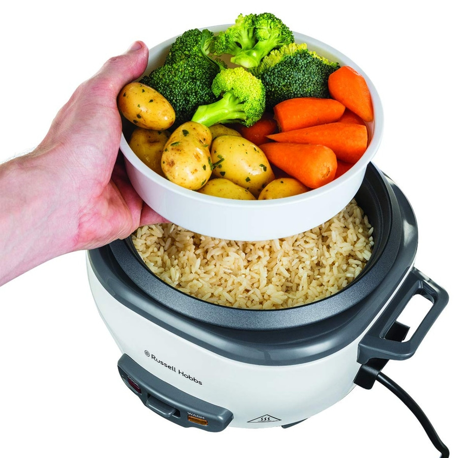 Eine Hand hält einen Dampfkorb mit Gemüse über einem Reiskocher mit Reis.