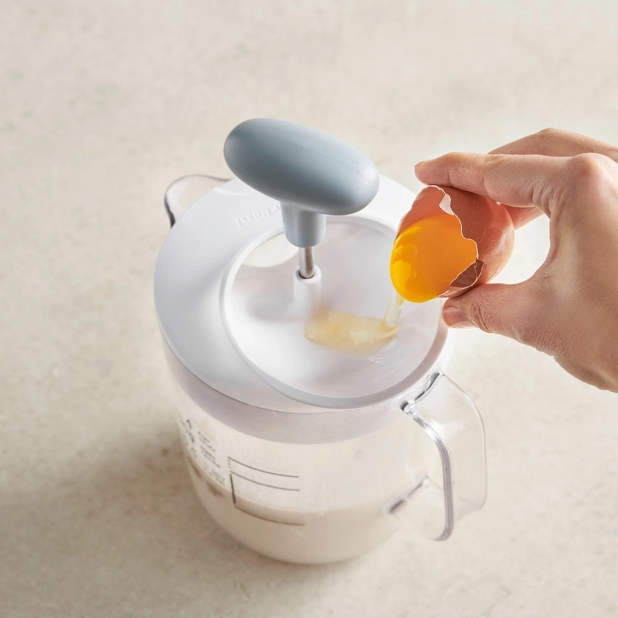 Hand hält Ei über Küchengerät, um es in eine Flüssigkeit zu gießen.