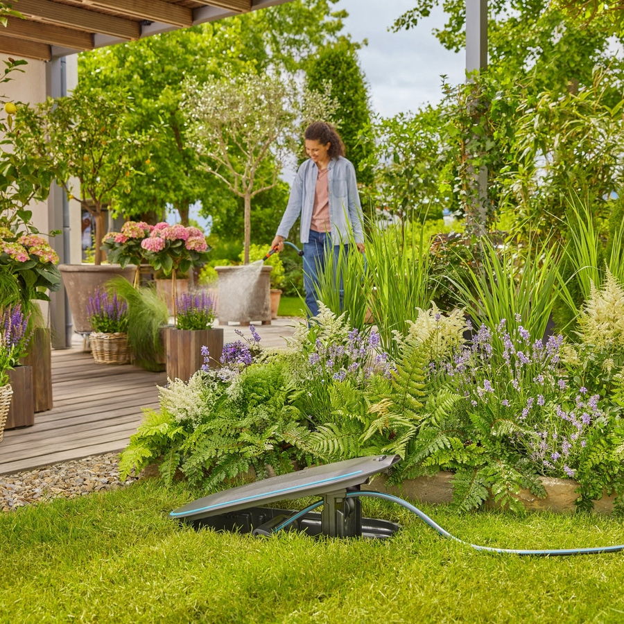 Eine Frau wässert einen Garten mit einem Schlauch, eine Bewässerungsanlage steht im Vordergrund.