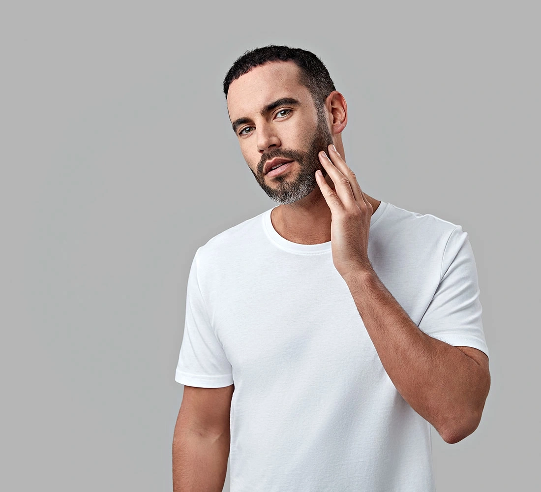 Mann mit Bart berührt sein Gesicht, trägt ein weißes T-Shirt