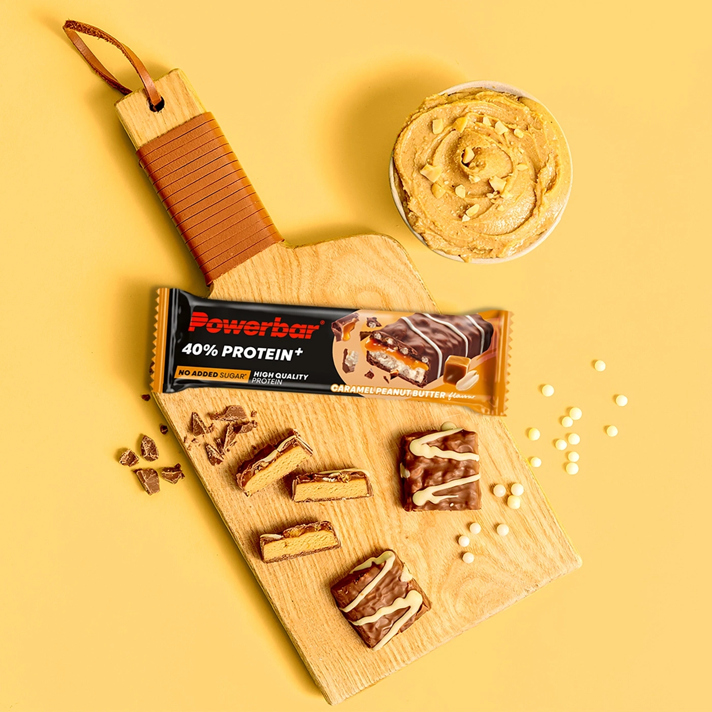 Powerbar Proteinriegel mit Karamell und Erdnussbutter-Geschmack, auf einem Holzbrett mit Erdnussbutter und Schokostückchen.