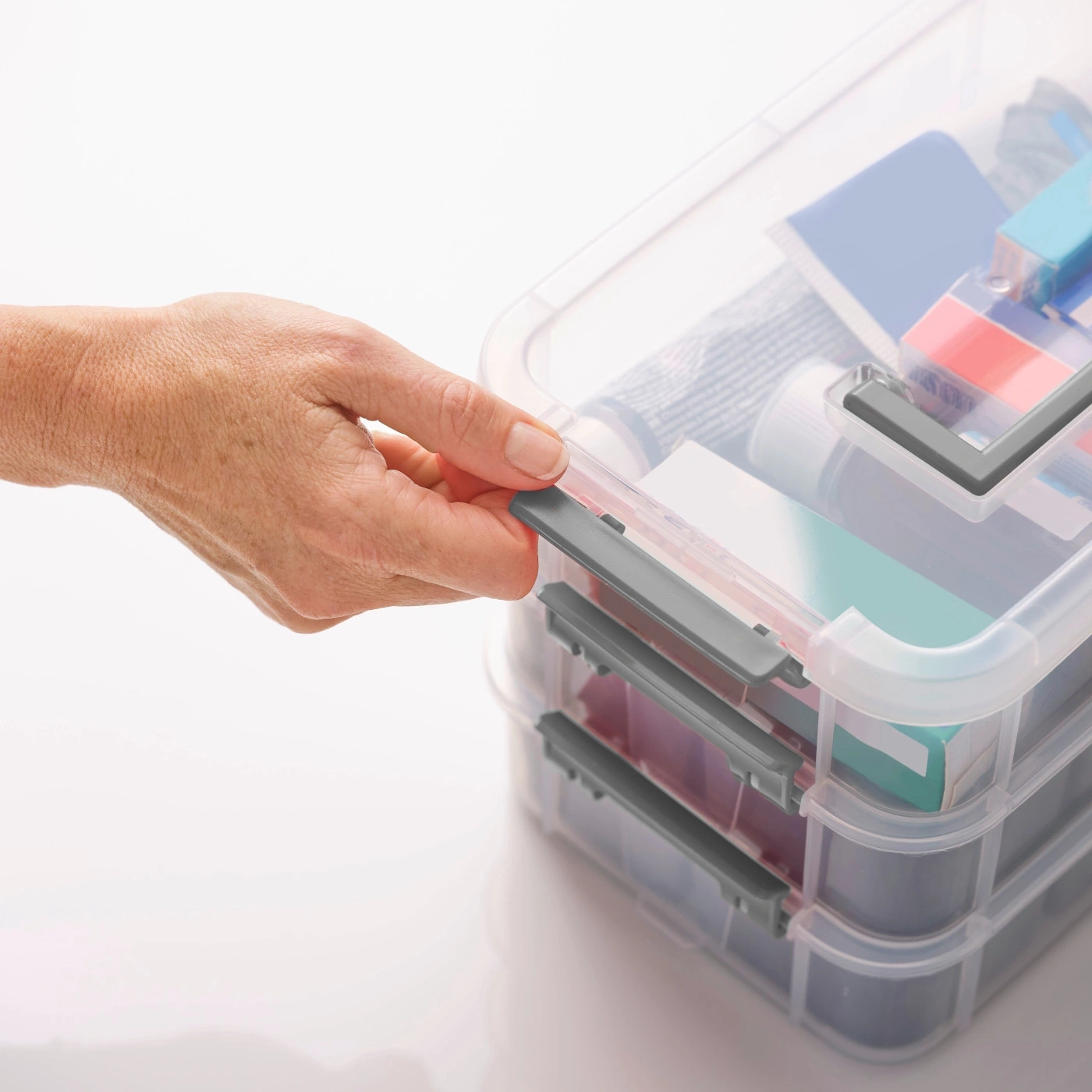 Hand öffnet eine transparente Aufbewahrungsbox mit grauem Verschluss. Mehrere Ebenen.
