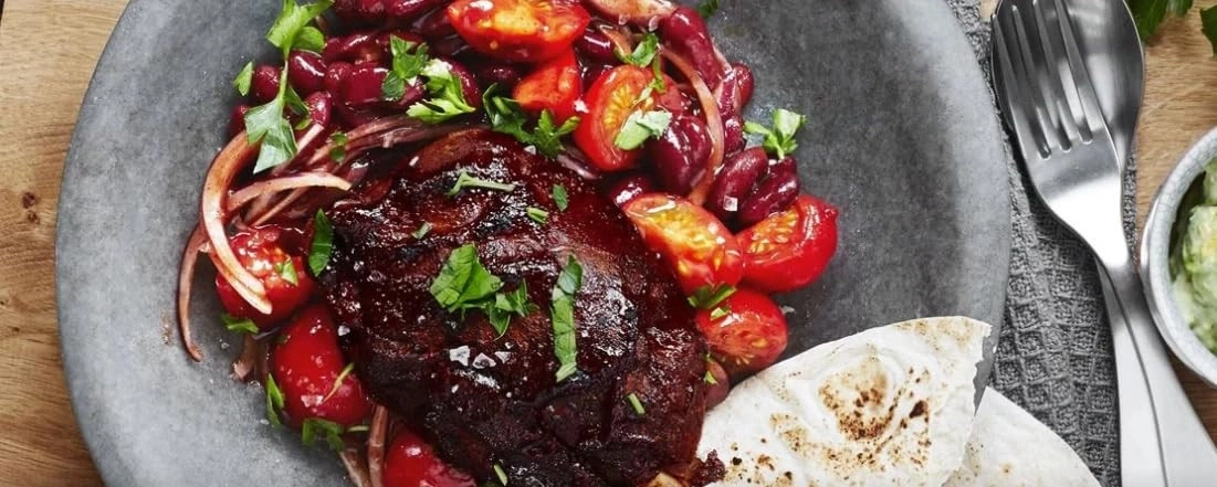 Ein Teller mit einem dunklen, glasierten Stück Fleisch, Kidneybohnen, Tomaten und Fladenbrot.